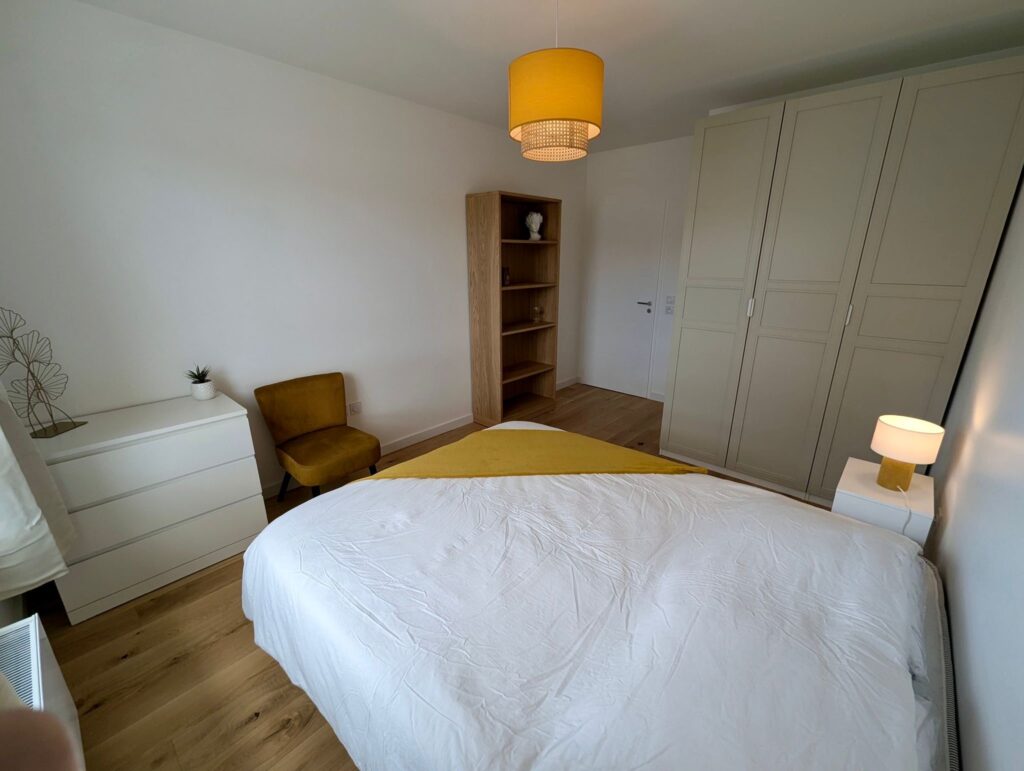 Chambre adulte confortable sur les tons jaunes avec un lit en 160 x 200 cm, une bibliothèque, un fauteuil carpeau et une penderie.